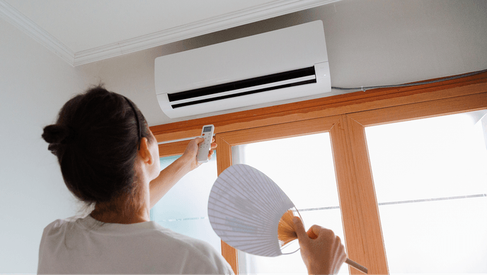 A woman waving herself with a hand fan while adjusting an air conditioner